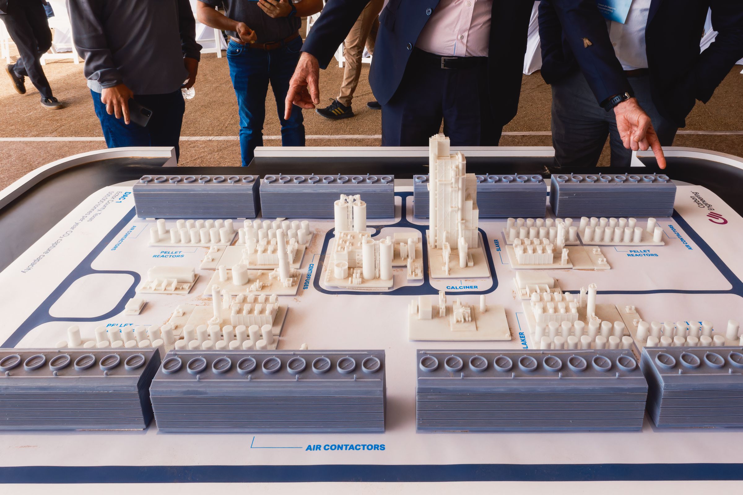 A closeup of several people standing behind and pointing to a small-scale model of a carbon removal plant.