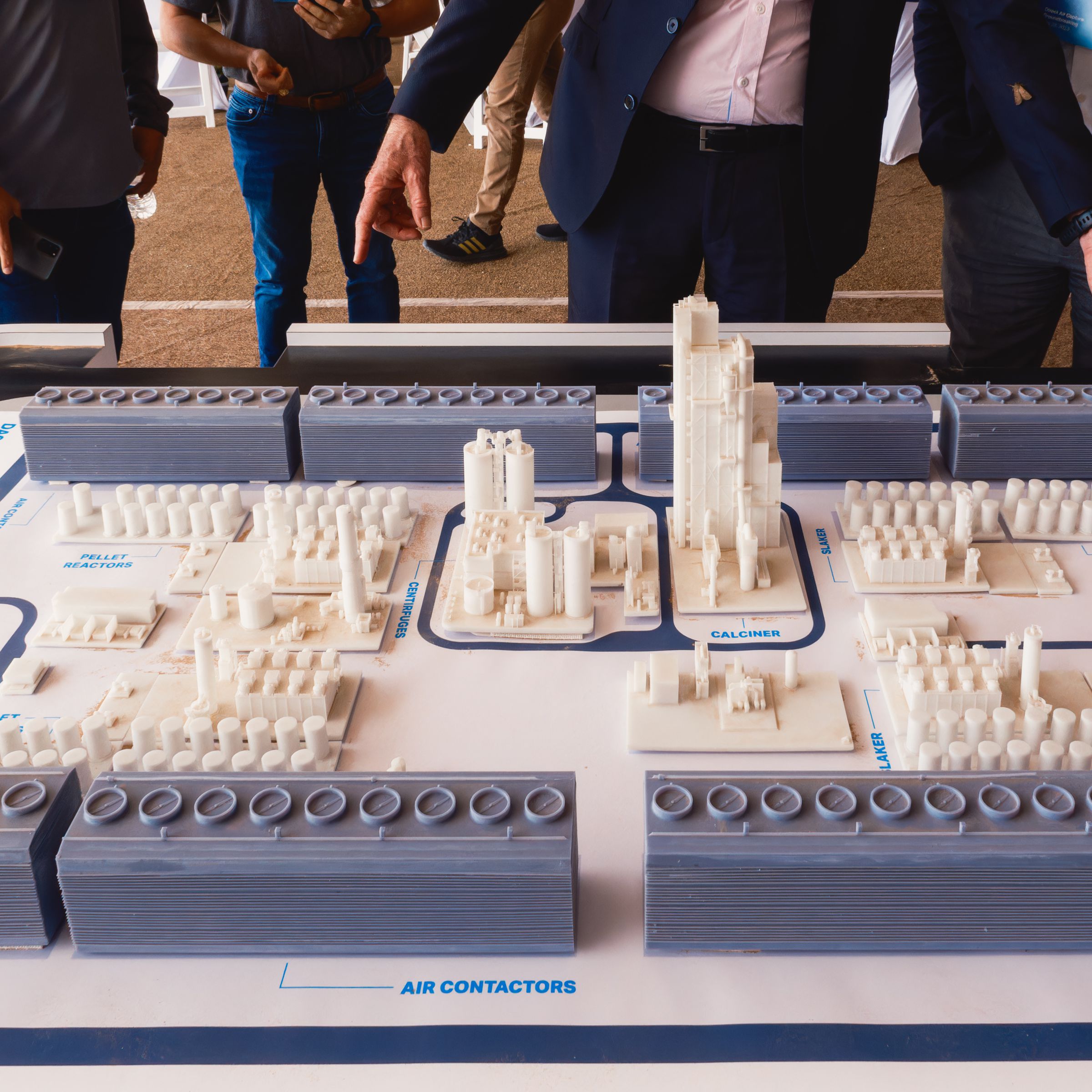 A closeup of several people standing behind and pointing to a small-scale model of a carbon removal plant.