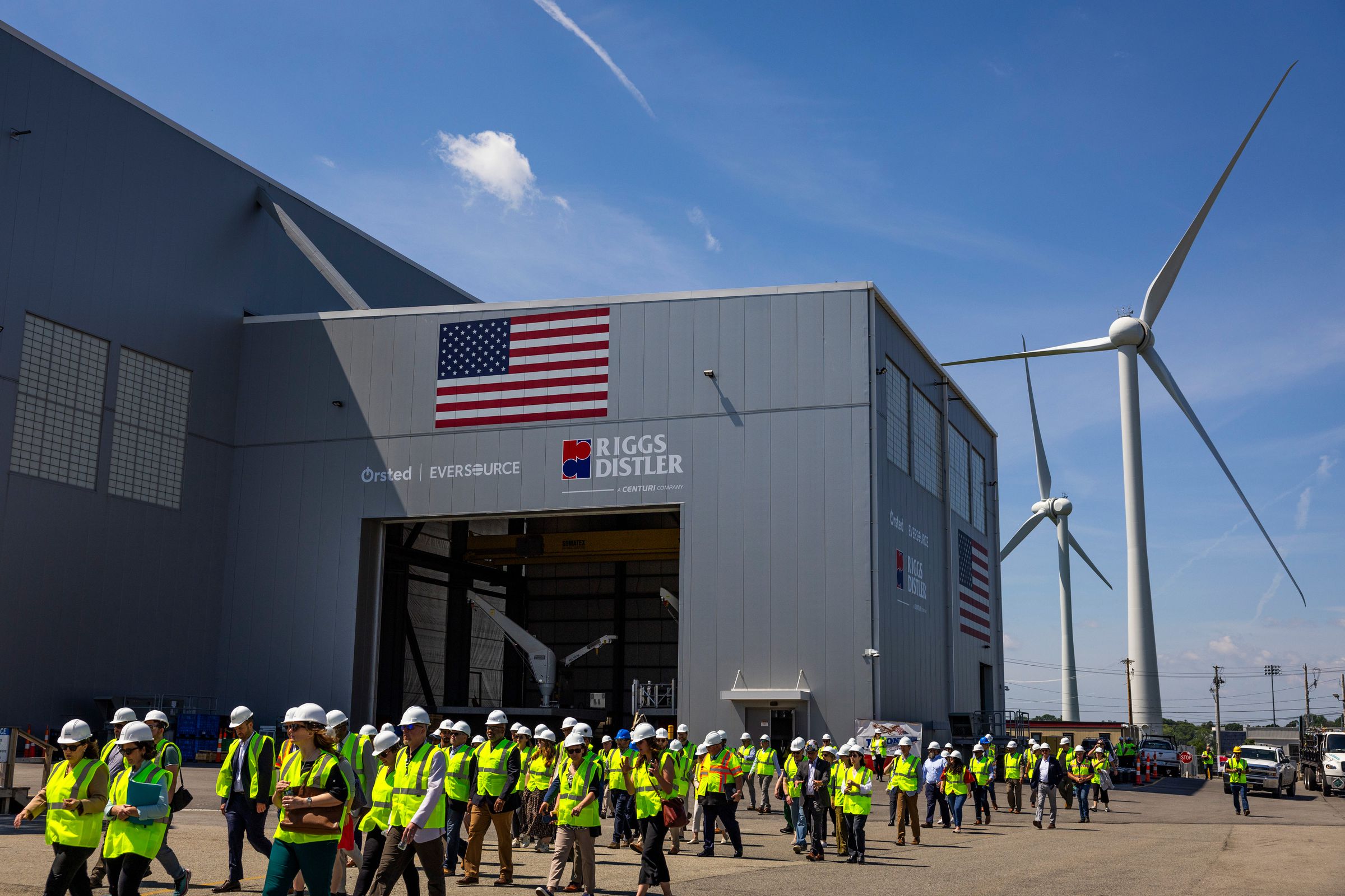 A line of people wearing construction vests and hard hats walk past a large industrial building with tall wind turbines seen behind them.