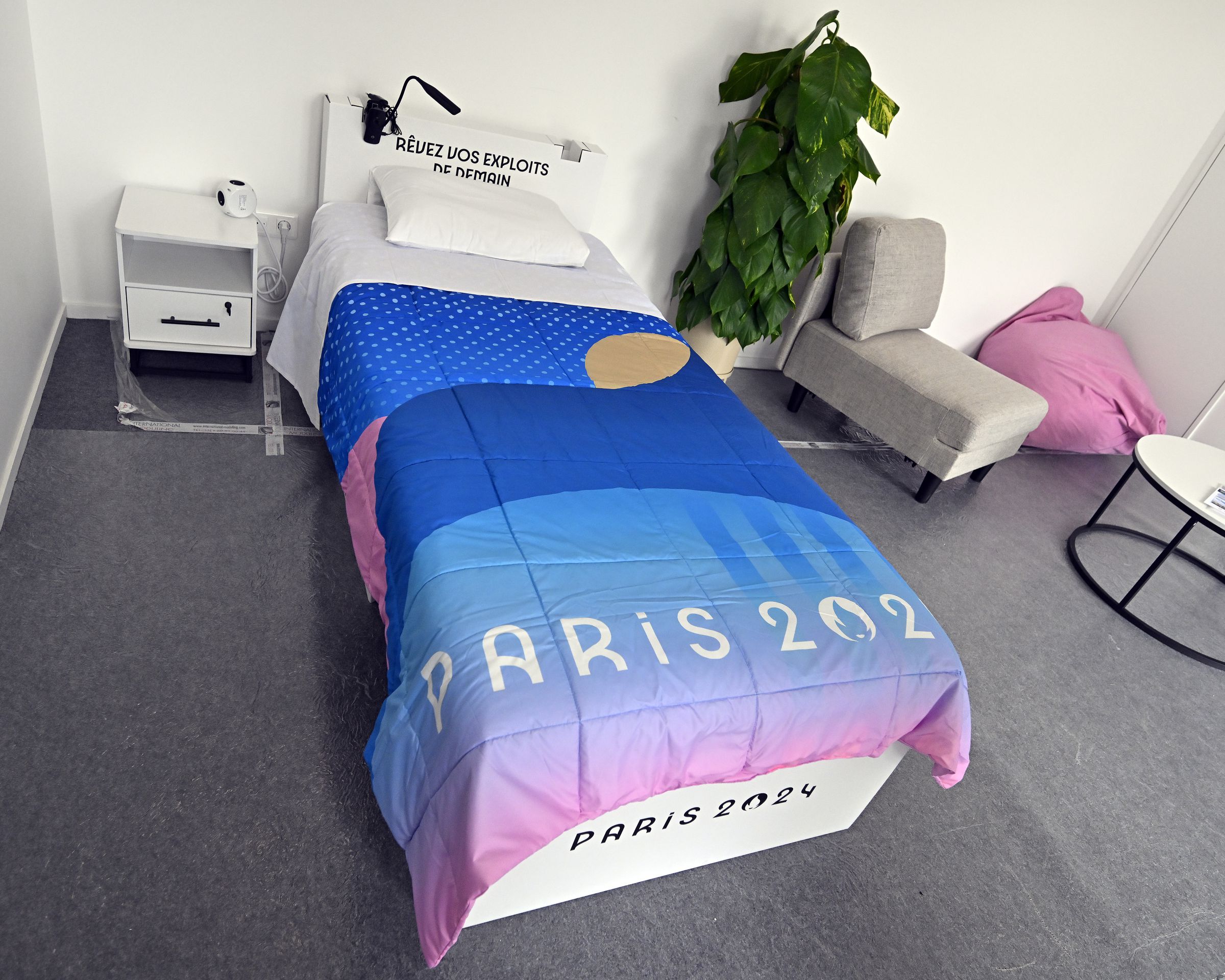 A room with a twin-sized bed covered with a blanket that says Paris 2024. There’s a nightstand on one side of the bed and on the other side a tall plant, a small lounge chair, and coffee table can be seen.