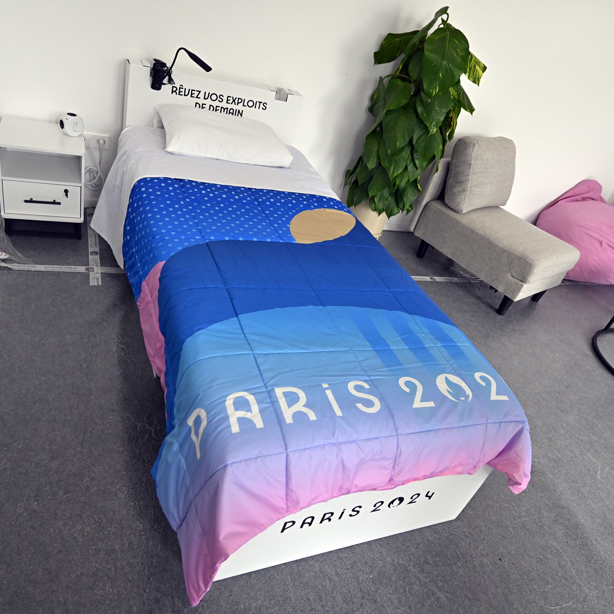 A room with a twin-sized bed covered with a blanket that says Paris 2024. There’s a nightstand on one side of the bed and on the other side a tall plant, a small lounge chair, and coffee table can be seen.