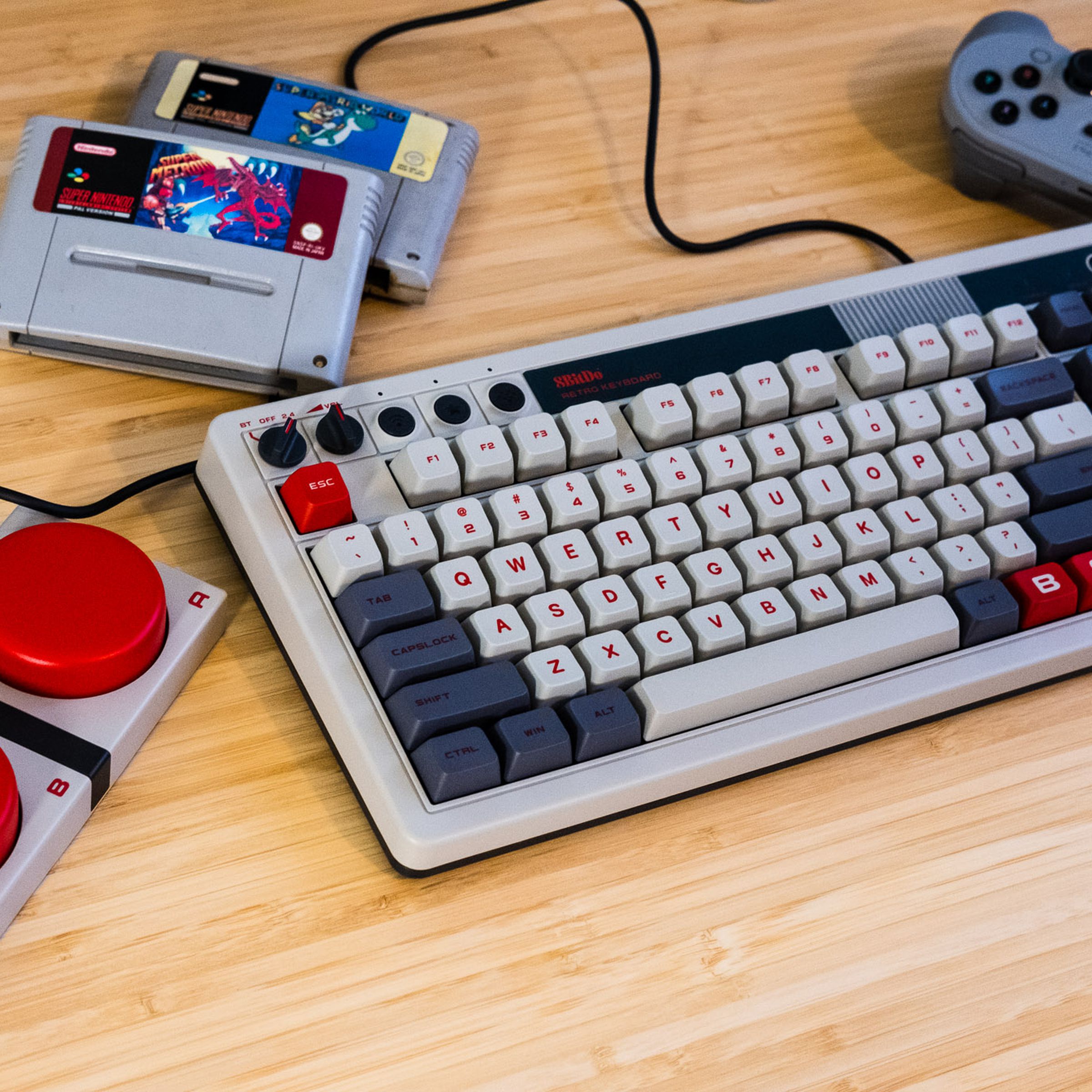 8BitDo Retro Keyboard on a desk next to NES-themed buttons, controller, and SNES cartridges.