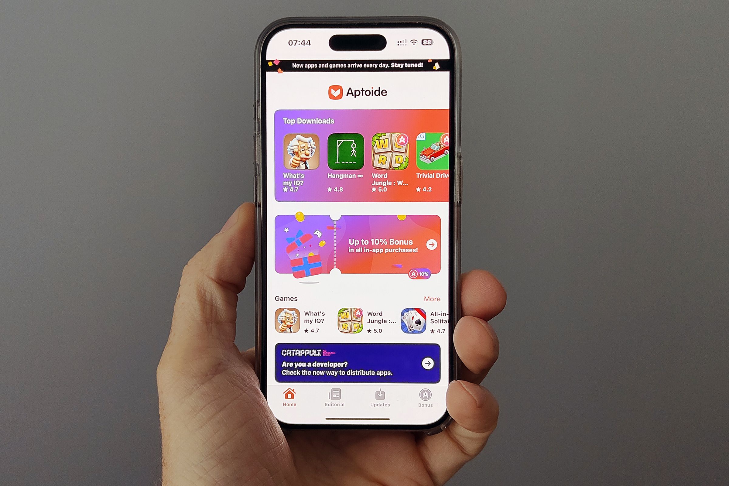 An iPhone held aloft in one hand shows the Aptoide iOS game store landing page full of colorful icons for the few games available.