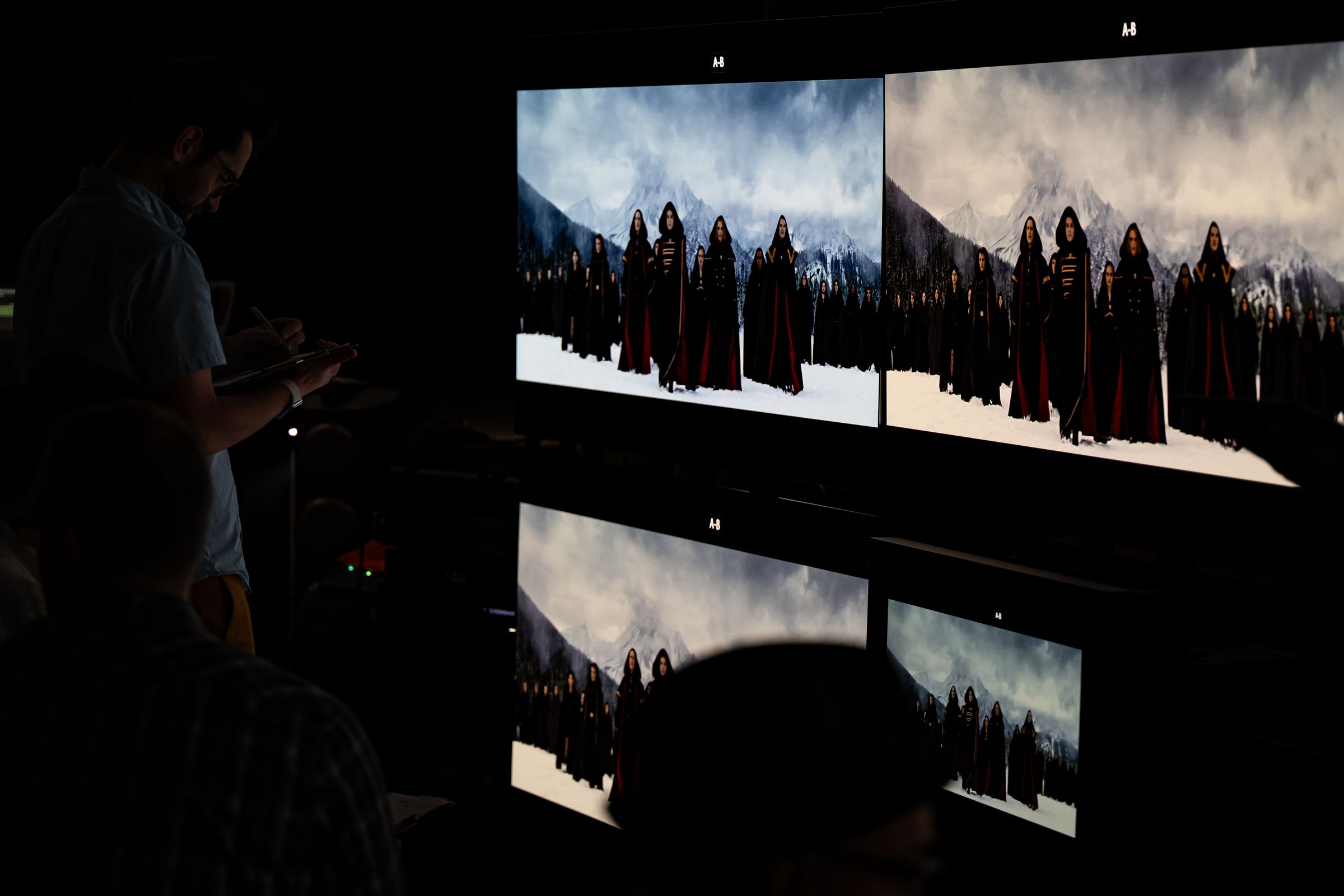 A group of large TVs and one small reference monitor in a dark room, showing a movie scene with hooded people against a cloudy sky.