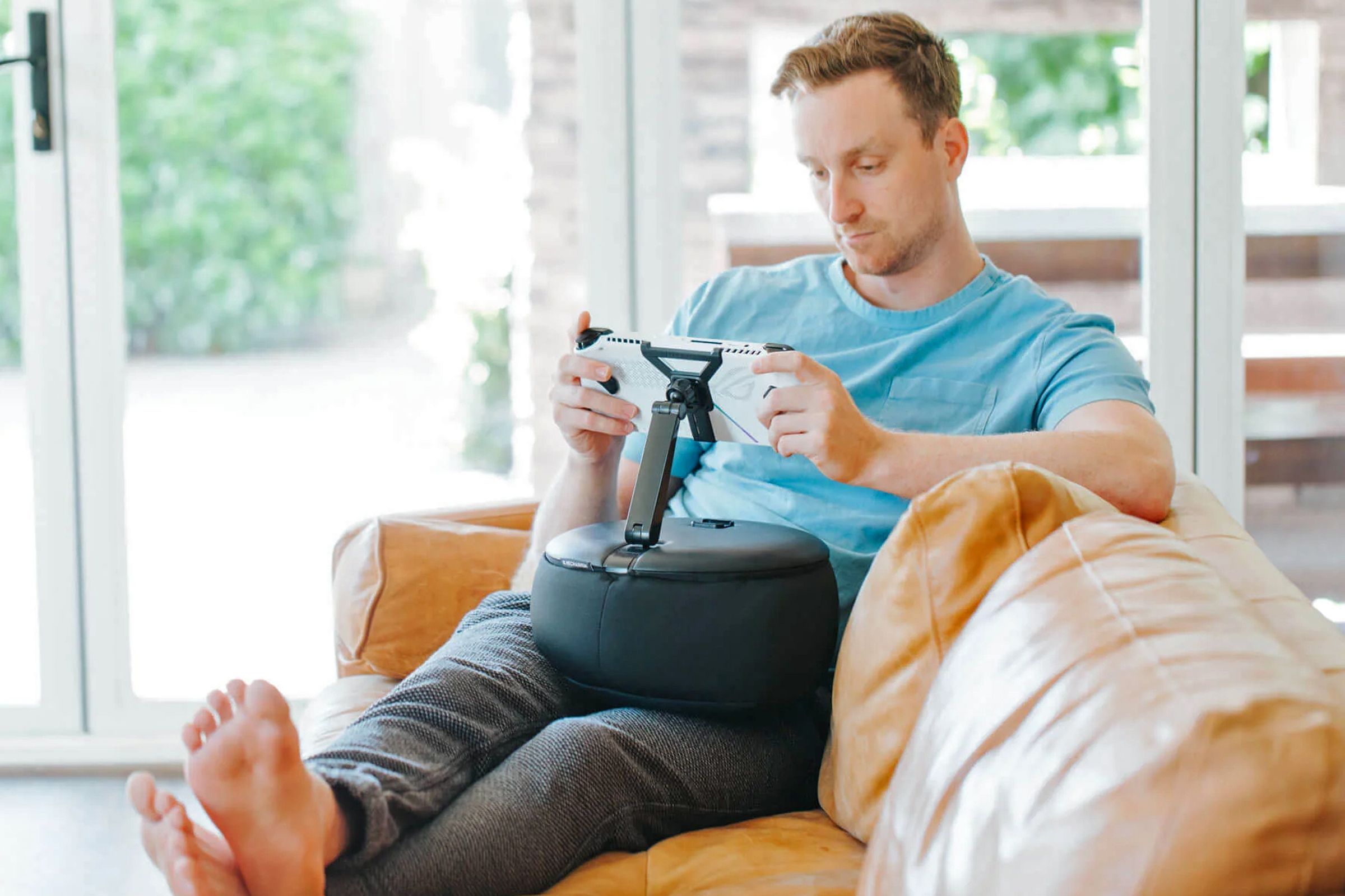 A gamer plays a handheld console on a sofa attached to the Gaming Pillow on their lap.