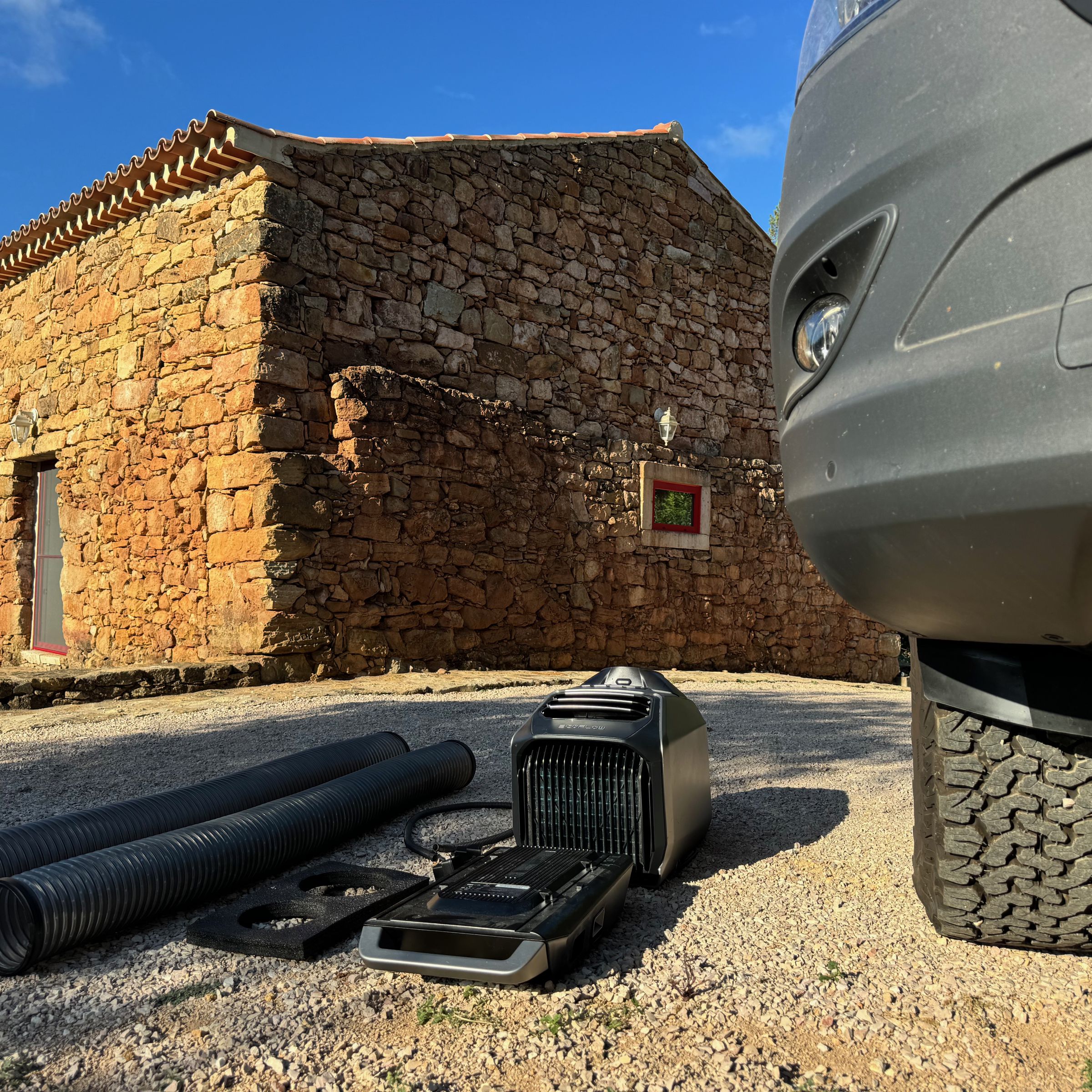 The Wave 2, its ducting, window vent board, drain and power cable sitting on gravel next to a large off-road wheel and an old stone farmhouse in the background.