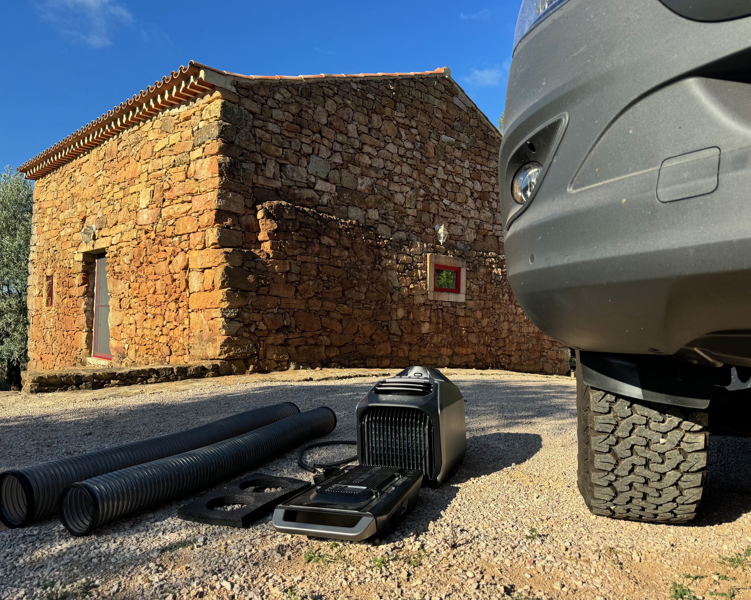 The Wave 2, its ducting, window vent board, drain and power cable sitting on gravel next to a large off-road wheel and an old stone farmhouse in the background.