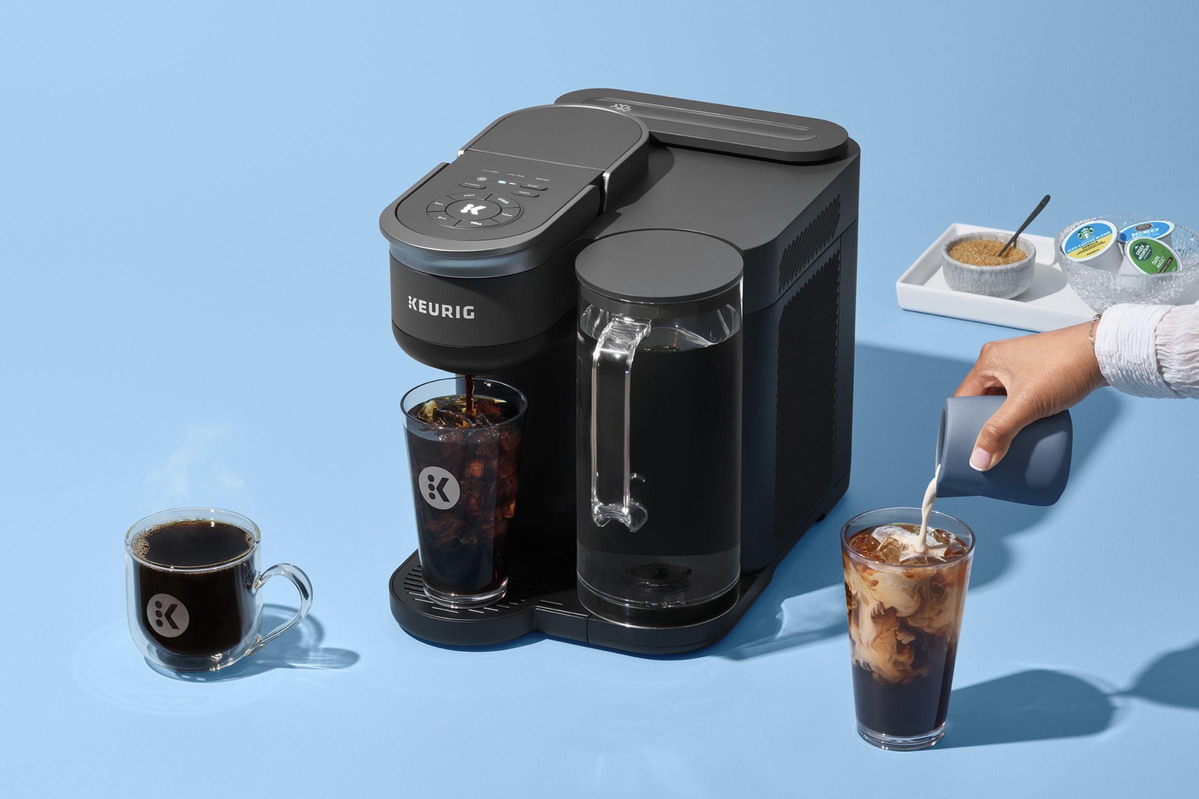 coffee machine with hot coffee cup, iced drink with hand pouring cream in, and sugar and coffee pods on a tray.