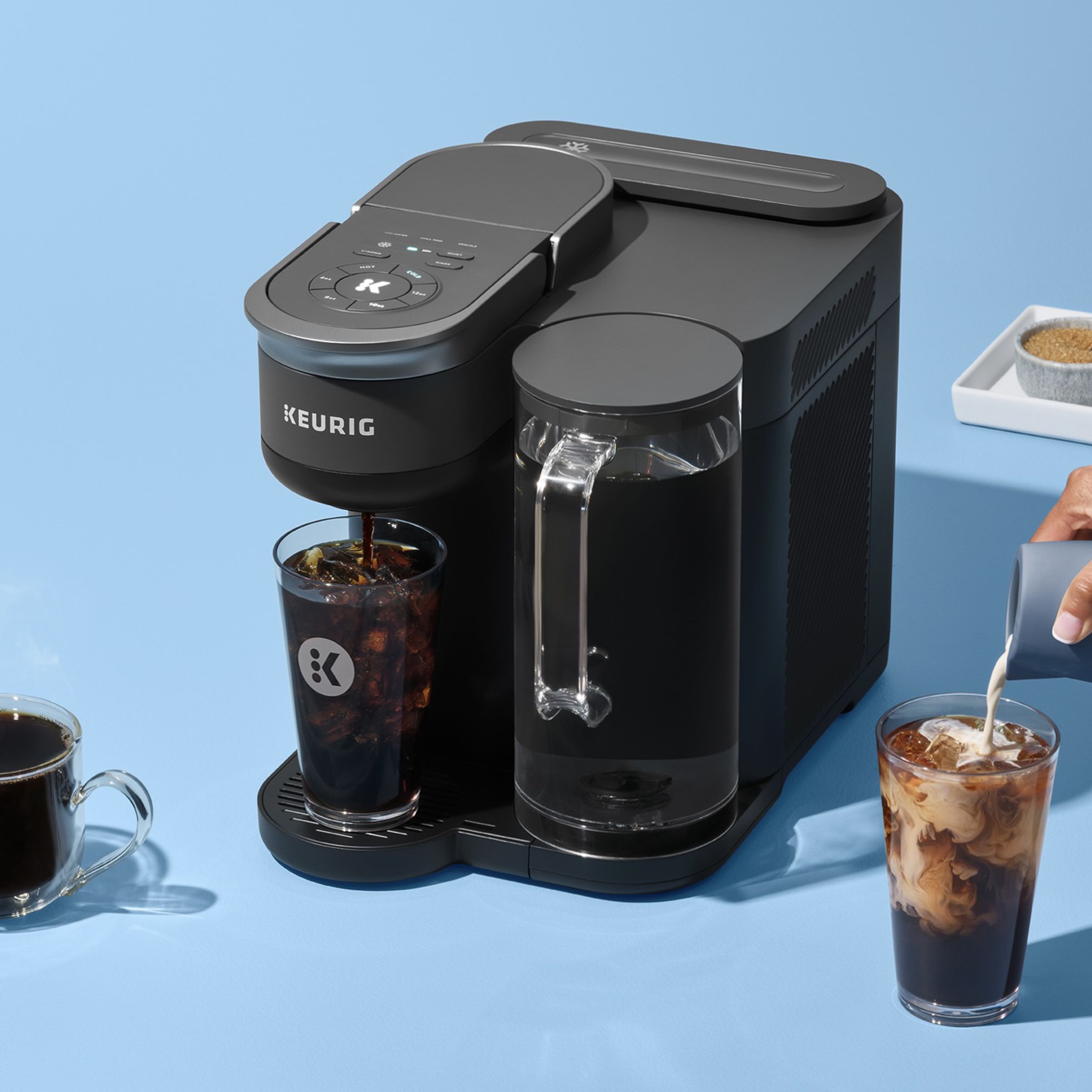 coffee machine with hot coffee cup, iced drink with hand pouring cream in, and sugar and coffee pods on a tray.