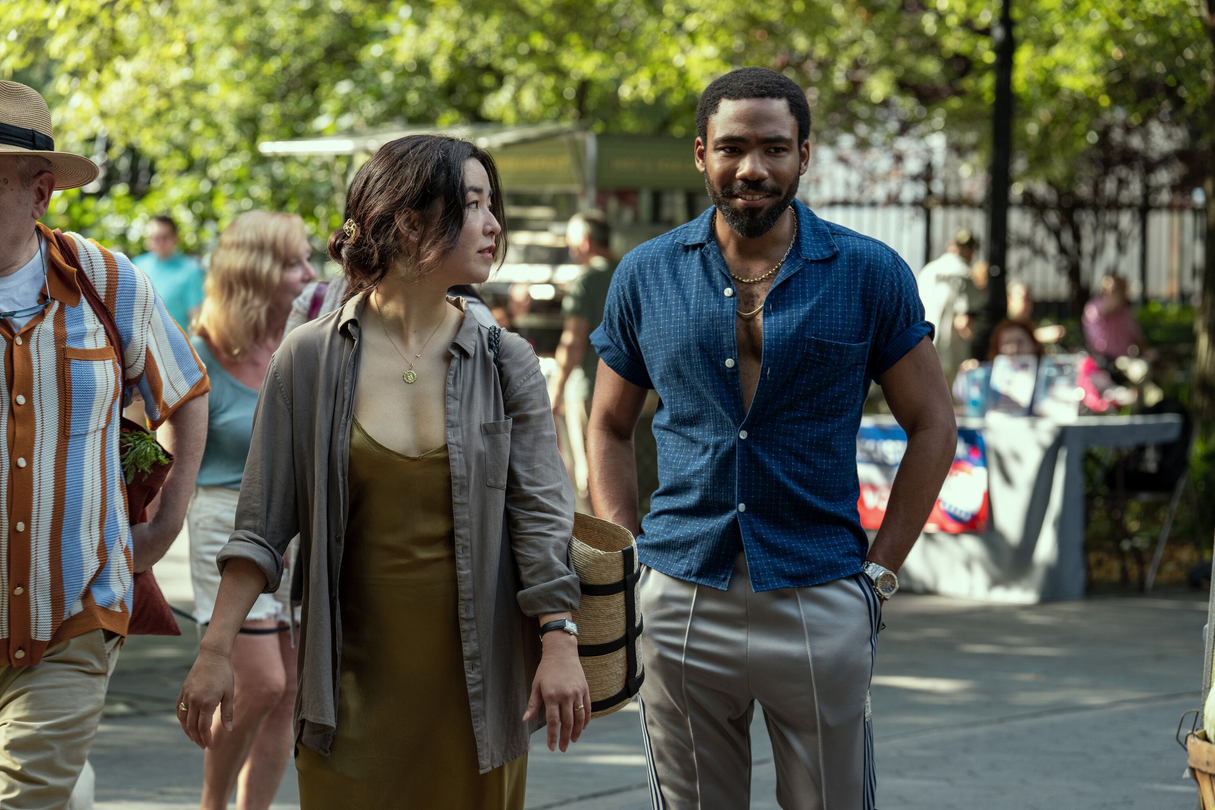 A woman wearing a low-cut brown dress and a gray collared shirt walking alonside a man in tan slacks and a short-sleeved blue shirt. The man and woman are walking through a park in spring. 