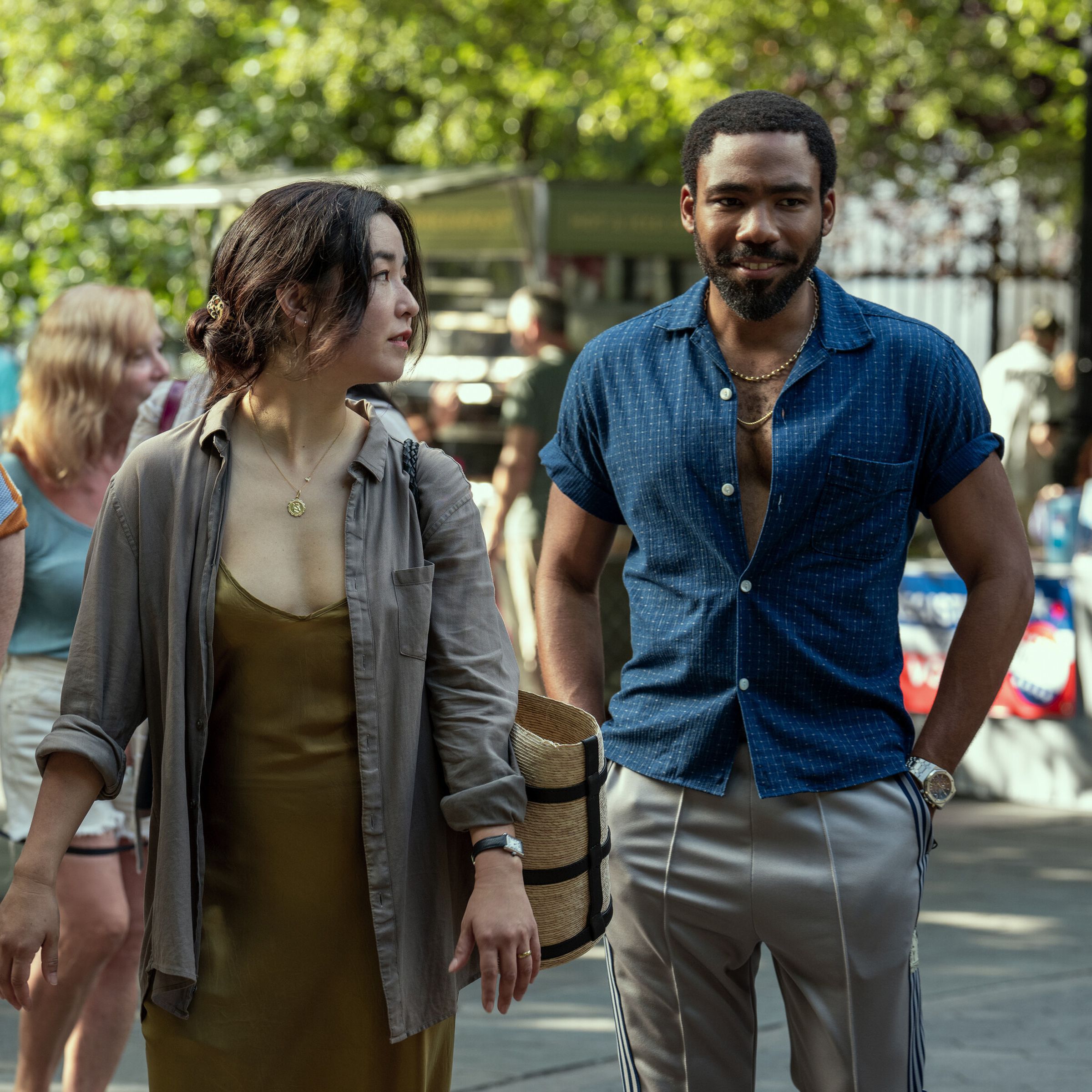 A woman wearing a low-cut brown dress and a gray collared shirt walking alonside a man in tan slacks and a short-sleeved blue shirt. The man and woman are walking through a park in spring. 