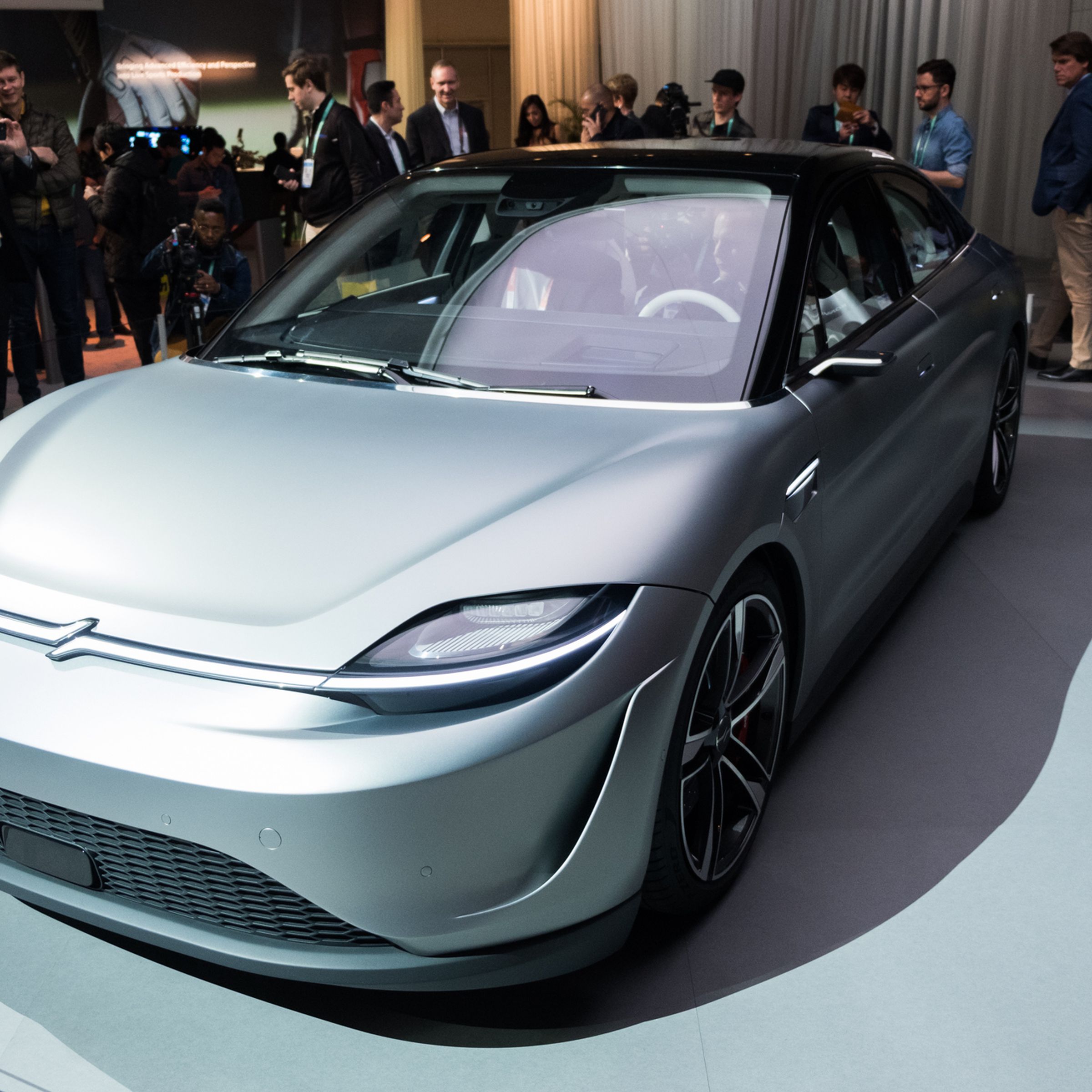 Sony’s Vision-S car is on the CES show floor with lots of people surrounding it. The car, a four-door midsize sedan, is silver with a black top and all-glass roof.