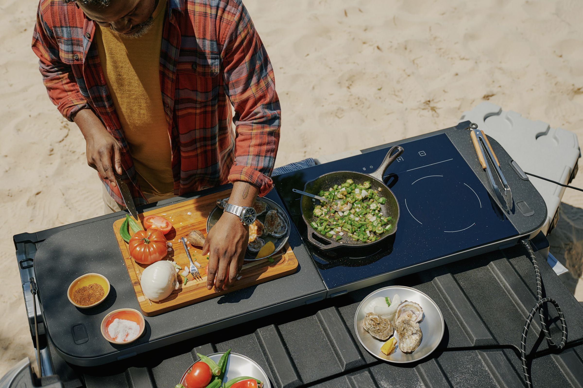 overhead view of the truck bed with a cooktop slab on top of the tailgate and a person cutting tomatoes on the cutting board side of the accessory with the induction stove frying veggies and a cable running to the side of the bed.