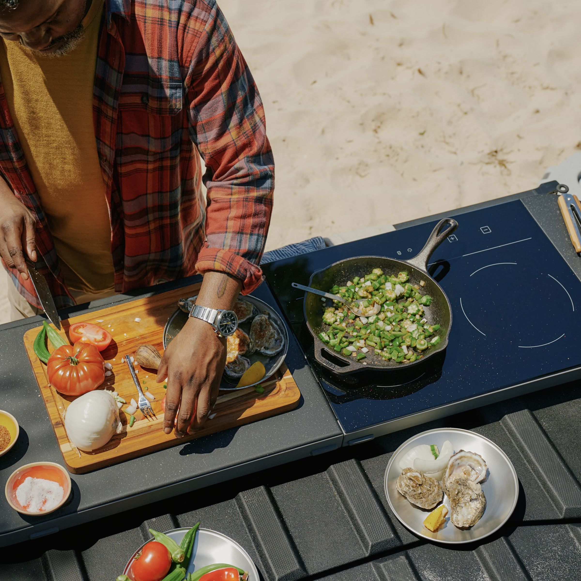 overhead view of the truck bed with a cooktop slab on top of the tailgate and a person cutting tomatoes on the cutting board side of the accessory with the induction stove frying veggies and a cable running to the side of the bed.